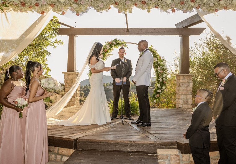 Bride reading vows beneath a floral pergola with the wedding party beside her